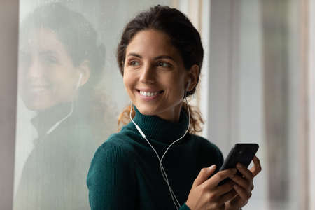 Pleasure to dream. Happy young latin female standing by window in earphones holding mobile phone dreaming by music song. Charming hispanic woman listen to audio book on cell look aside with smile.の写真素材