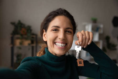 Selfie of happy homeowner. Excited latin female take self portrait on phone proud of buying house of dream paying mortgage. Young lady lottery winner look at camera shoot picture with keys of new flatの写真素材