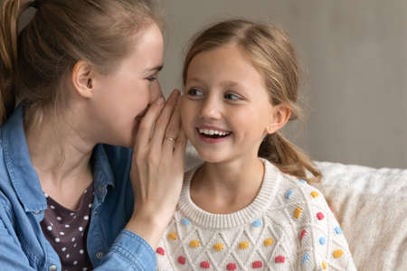 Young Caucasian mother telling secret whispering on ear of curious excited small adorable child daughter, gossiping or sharing confidential information at home, enjoying trustful conversation.の写真素材