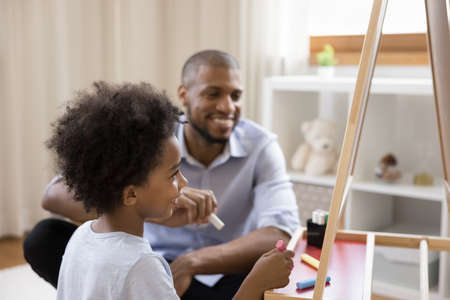 Happy cute Black little kid and dad drawing on chalkboard at home, using colorful chalks and whiteboard, laughing, having fun, enjoying leisure, playtime, entertainment, craft creative hobbyの写真素材