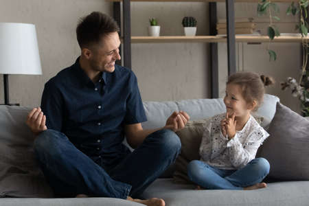 Happy dad teaching cheerful preschool daughter to meditate at home, sitting on couch with zen hands, talking to child, laughing. Daddy and kid doing exercise for mental health, mindfulnessの写真素材