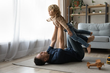 Focused strong daddy playing active sportive games with preschool son, lying on heating carpeted floor with toys, holding boy with flying hands for making airplane, doing acroyoga baseの写真素材