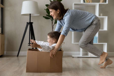 Happy active mom laughing, dragging box with screaming little daughter kid on floor at home. Mother and preschool child celebrating moving into new house, enjoying funny relocation partyの写真素材