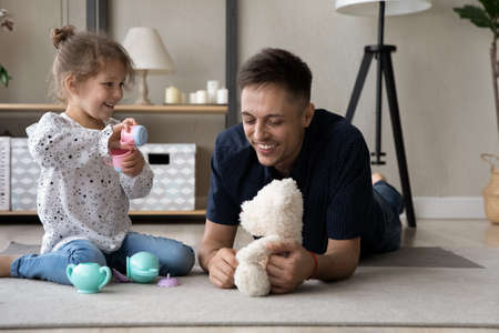 Happy laughing caring dad and daughter kid playing together on heating floor, relaxing in living room, having fun at tea party with teddy bear, toy cups, pot, dish. Fatherhood, family playtimeの写真素材