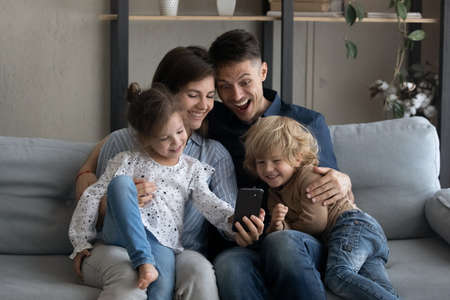 Cheerful daughter girl taking selfie on smartphone with beloved family. Happy couple of parents and two sibling kids relaxing on sofa, posing for self portrait, hugging, smiling, laughing on couchの写真素材