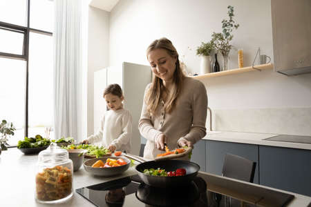 Adorable happy little preschool kid girl chopping fresh vegetables while smiling caring young mother putting it in pan for frying, enjoying cooking together in modern kitchen, healthy dieting concept.の写真素材