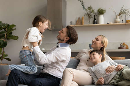 Bonding relaxed happy young mother and small son watching joyful father lifting in air laughing 6s preschool kid daughter, cheerful multigenerational family having fun together on weekend at home.の写真素材