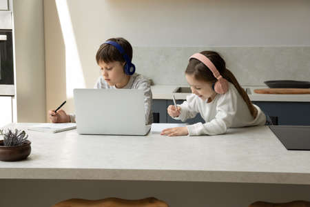 Happy little children siblings wearing wireless headphones watching educational lecture on computer or attending online lesson with teacher, writing notes in copybook, studying distantly at home.の写真素材
