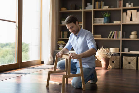 Happy homeowner man assembling wooden furniture at new home. Positive renter, tenant doing carpenter work on floor, constructing chair, putting together pieces of rack. DIY, housing conceptの写真素材