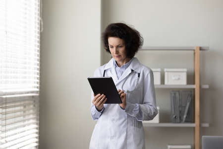 Serious mature female doctor using tablet computer. Elder practitioner giving online virtual consultation, chatting to patient, making appointment on internet medical app, reading electronic recordsの写真素材