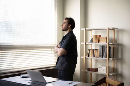 Serious thoughtful business man thinking over company future in office, standing at workplace, looking out of window with folded hands, making decision, pondering over startup project issuesの写真素材