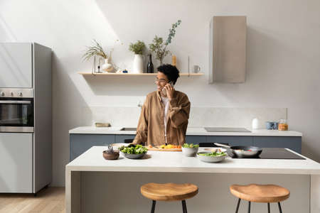 Happy young attractive African American woman in glasses holding pleasant cellphone call conversation, communicating with friends inviting for domestic party, distracted from preparing food in kitchenの写真素材