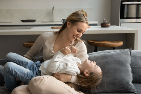 Happy young 35s Caucasian mother tickling joyful cute little 6s child daughter, having fun playing...