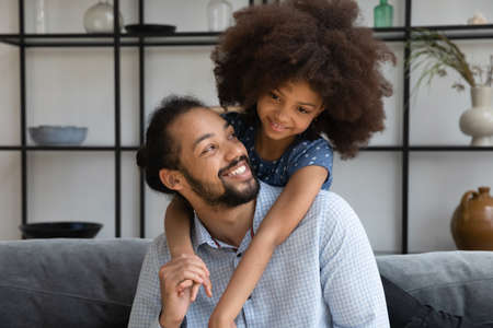 Cheerful small African American kid girl cuddling from back sincere father, enjoying communicating at home, showing sweet tender emotions, enjoying peaceful carefree moment together at home.の写真素材