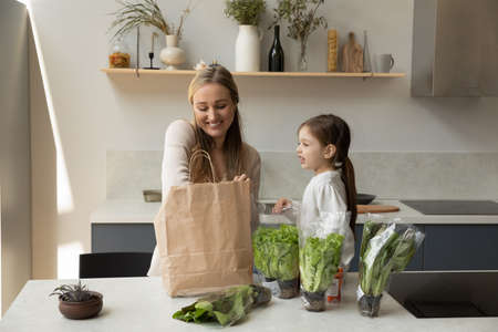 Happy young mother and joyful cute small kid daughter unpacking fresh salad leaves and organic greenery from carton package, unpacking products for healthcare eating together in modern kitchen.の写真素材