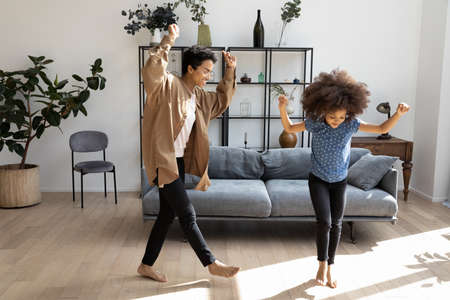 Full length overjoyed millennial African American mother or babysitter dancing to favorite energetic disco music with happy small adorable child barefoot on warm wooden floor in modern living room.の写真素材