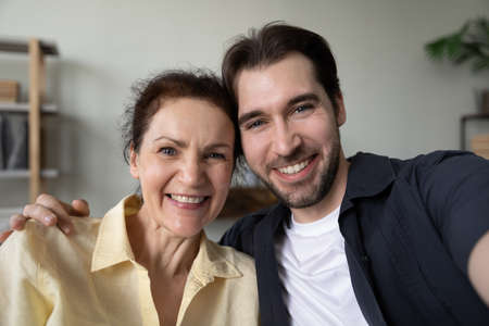 Happy adult son and mature mom hugging with love, affection, taking selfie, looking, smiling at camera, having fun, enjoying leisure at home together. Head shot portrait. Motherhood conceptの写真素材