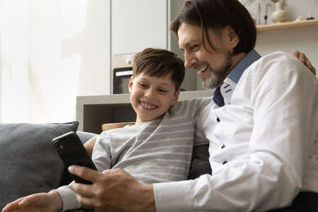 Joyful two male generations family looking at smartphone screen, enjoying using funny mobile software application, laughing on entertaining photo video online in social networks or shopping at home.の写真素材