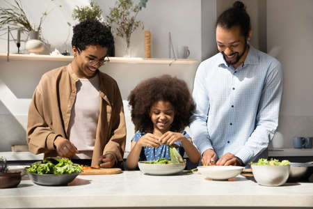 Joyful two generations African American family preparing healthy food, cutting fresh organic vegetables on wooden board. Happy multiracial couple parents cooking with little child daughter in kitchen.の写真素材