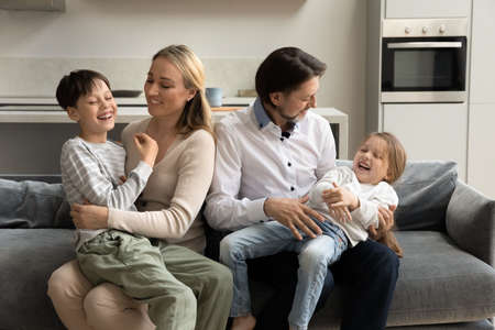 Happy loving young couple parents tickling adorable small preschool son daughter children, enjoying entertaining family activity, having fun together resting on comfortable couch at home on weekend.の写真素材