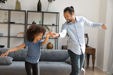 Happy little African American kid girl dancing to energetic disco music with caring smiling father in modern living room, joyful two generations family enjoying hobby activity together on weekend.の写真素材