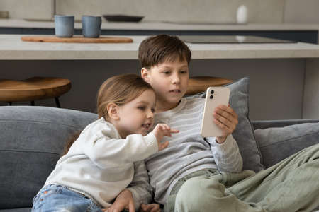 Addicted to modern tech happy small adorable children siblings using cellphone software applications, playing entertaining mobile games, watching funny videos, spending time online, sitting on couch.の写真素材