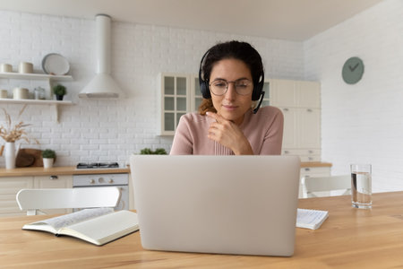 Focused woman in headset looking at laptop screen, studying online, sitting at table in modern...