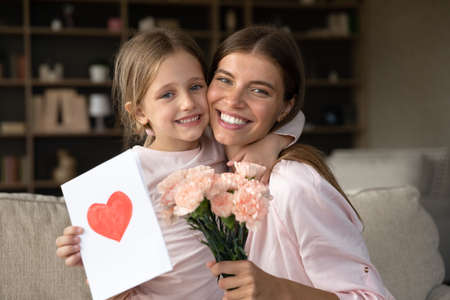 Grateful mom hugging daughter girl, holding flowers bouquet, receiving hand drawn greeting card with...