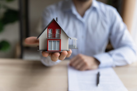 Close up realtor sit at table holds tiny house model, layout of cottage. Real estate purchase, new property, ownership, construction company make special offer to client, affordable dwelling conceptの写真素材