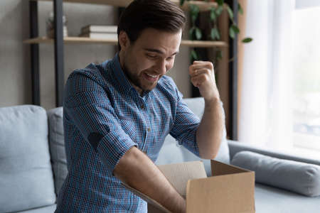 Young man sit on sofa at home opens cardboard parcel box with long-awaited goods feels overjoyed...