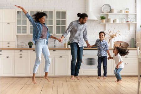 Beautiful African couple and little cute children holding hands jumping barefoot together in fashionable kitchen on warm wooden floor. Active weekend, new modern house, happy homeowners family conceptの写真素材