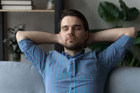 Front view young serene man put hands behind head falls asleep, takes day nap, resting leaned on cozy soft sofa in living room with air conditioning system. No stress, modern house, relaxation conceptの写真素材