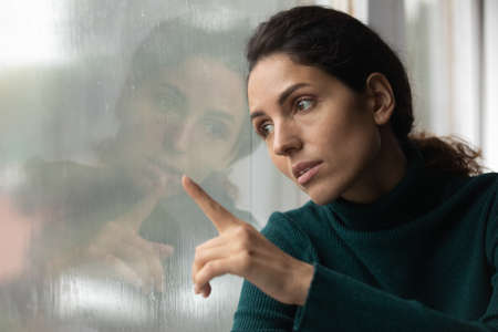Upset young Latino woman look in window distance feel depressed unhappy alone at home. Sad stressed millennial Hispanic female on lockdown indoors, lack communication. Solitude, loneliness concept.の写真素材