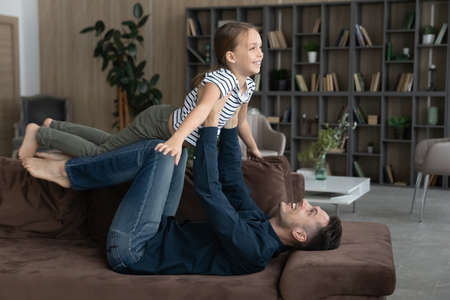 Happy young father lifting in air adorable little child daughter, lying on comfortable couch at home. Happy two generations family having fun, imagining travelling, playing airplane game on weekend.の写真素材