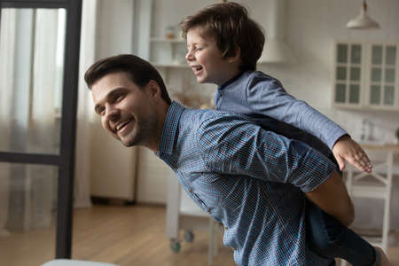 Loving Caucasian father piggy backing laughing little son, smiling caring young Caucasian dad carrying adorable 5s child boy, family engaged in funny activity, enjoying leisure time at home togetherの写真素材