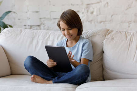 Happy small teenage child boy holding digital computer tablet, having fun online playing games or...