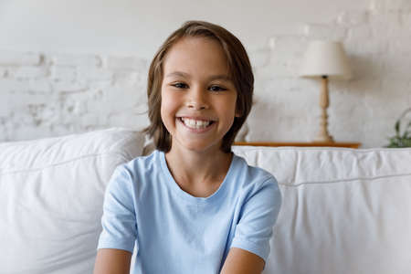 Head shot portrait of adorable small preteen kid boy with long hairstyle sitting on couch. Smiling little 12s child looking at camera, enjoying distant video call distant conversation at home.の写真素材