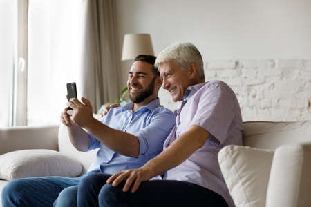 Happy two male generations family using cellphone, posing for funny selfie photo, recording video or...