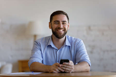 Portrait of smiling young handsome businessman manager employee holding smartphone in hands, sitting at table in home office. Addicted to modern technology millennial man using cellphone software appsの写真素材