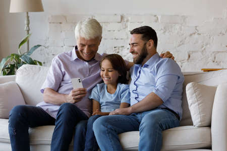 Happy middle aged old man holding cellphone in hands, posing for selfie photo with laughing adorable preteen grandchild and grownup son, enjoying pleasant distant video call or watching funny content.の写真素材
