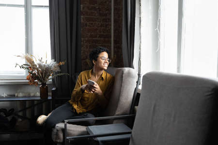 Dreamy young African American woman in glasses looking out of window in distance, holding cellphone...