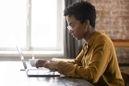 Side view focused young African American businesswoman in eyewear working on computer, sitting at...