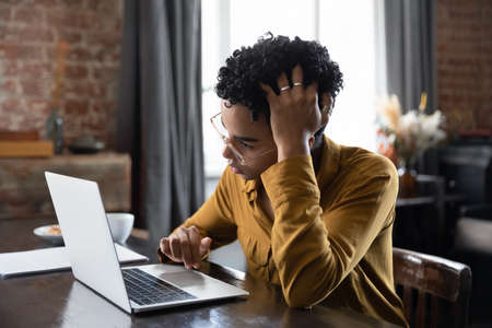 Unhappy confused millennial African American woman in glasses looking at laptop screen, stuck with...