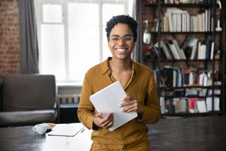 Portrait of joyful young African American woman in eyeglasses holding digital computer tablet in hands, posing in modern loft style workplace, feeling motivated, modern tech usage for business.の写真素材