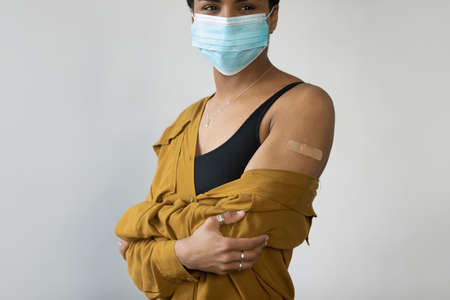 Cropped young pretty African American woman wearing protective medical facemask, showing place on arm with glued patch after coronavirus vaccination injection, isolated on white studio background.の写真素材