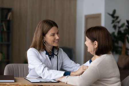 Cardiology checkup. Friendly young female doctor family therapist examine heart lungs of senior woman patient using stethoscope. Old age lady visit cardiologist to check cardio health get consultationの写真素材