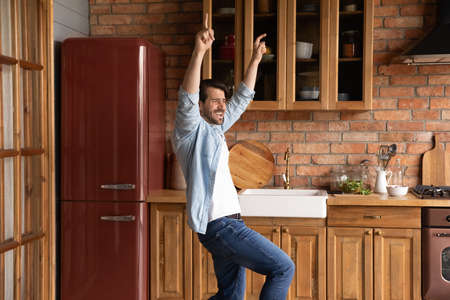 Overjoyed funky handsome young male homeowner singing song, dancing to disco music in old fashioned kitchen, enjoying stress free leisure weekend pastime alone at home, domestic activity concept.の写真素材