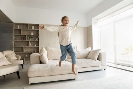 Overjoyed energetic happy little child girl jumping from comfortable couch on floor, having fun alone in modern light living room, involved in entertaining funny active game on weekend at home.の写真素材