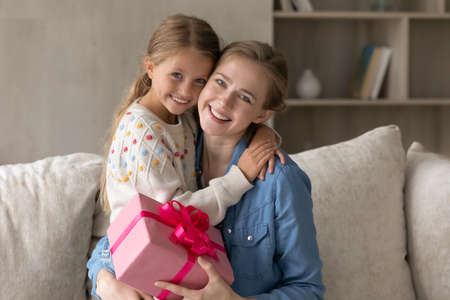Portrait of happy bonding two female generations family holding wrapped gift box in hands. Joyful loving young mother cuddling cute little child daughter, feeling thankful for prepared gift at home.の写真素材