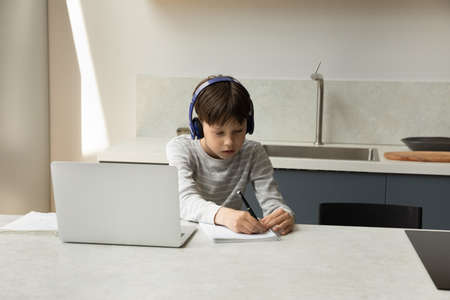 Focused gen Z teen school boy in headphones attending virtual class, listening to teachers audio...